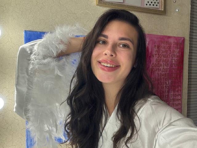 BlackFatal, con lunghi capelli scuri, sorridente, indossa un morbido accappatoio bianco, con una mano dietro la testa, rossetto rosa, seno di media grandezza, pelle chiara, occhi marroni, in una stanza luminosa..