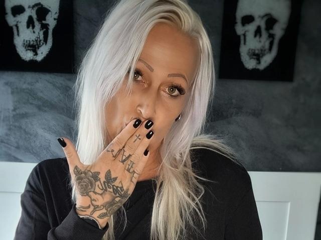 Hotjassy-hot with platinum blonde hair, black nail polish, tattoos on hand, covering mouth, green eyes, black shirt, skull paintings in background, looking at camera, serious expression.