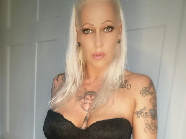 Hotjassy-hot with platinum blonde hair, black lace bustier, tattoos on shoulders and chest, large tits, pierced ears, and full pink lips, staring into the camera with a serious look.