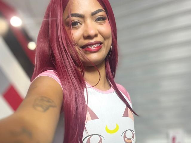 ChloeBrownOne has red hair, a smile with visible teeth, pink shirt with cat eyes, and a tattoo on her right arm. Her pink shirt has a crescent moon.