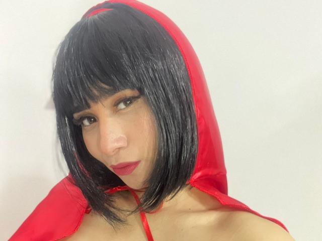 MelannyTaylorr with shiny black bob, red hood, and red lipstick, wearing a red top, looking sideways with a slight smile. Boobs visible, red top straps on shoulders.