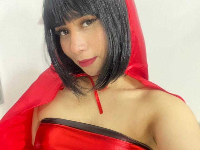 MelannyTaylorr with short black hair, red dress, and matching hood, showing off her tits, red lipstick, and subtle makeup. She's looking at the camera with a slight smile.