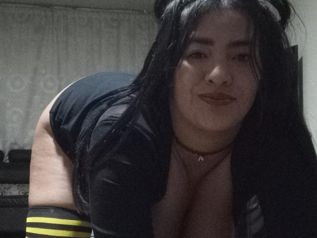 ArianithaXox with long black hair, wearing a black top and shorts, bending forward, showing large tits and a plump ass, smiling, in a dimly lit room.