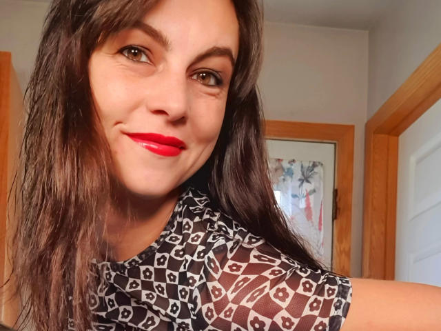 LillyMania-hot with wet brown hair, red lipstick, and a patterned black shirt, smiling at the camera, showing off her cute face and small tits.