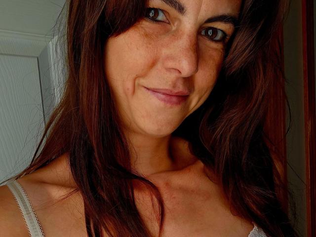 LillyMania-hot with brown wavy hair, freckles, and a subtle smile, wearing a white spaghetti strap top, showing a small bruise on her chest, brown eyes, and a cute face.