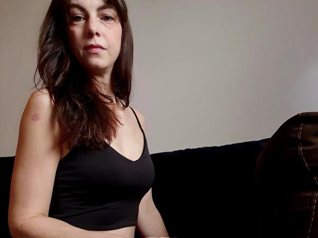 LillyMania-hot with dark brown hair, wearing a black sports bra, showing a small red bruise on her right shoulder, sitting on a black couch, looking at the camera.