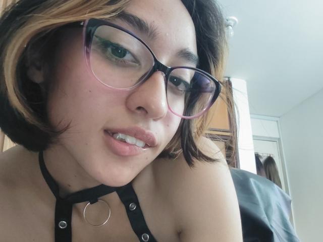 CamiVargas with short brown and blonde hair, pink glasses, black halter with ring, slightly parted lips showing teeth, green eyeshadow, and a cute, sexy expression.