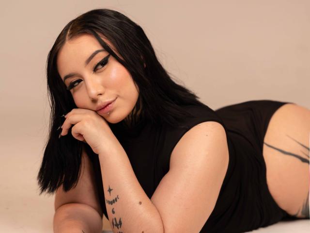 NinaRaven with black hair, lying on her stomach, wearing a black sleeveless top, showing tattoos on her arm and side, looking at the camera with a slight smile.