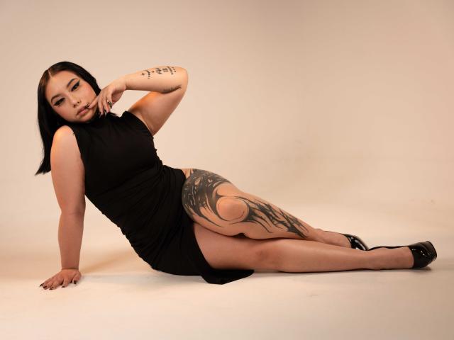 NinaRaven is a curvy Asian woman with black hair, wearing a black dress, showing a large tattoo on her right thigh, and black heels, lying on a white floor.