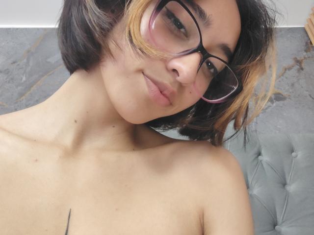 CamiVargas mit kurzen, schwarzen und blonden Haaren, die rosafarbene Brille trägt, lächelt, zeigt ihre nackten Schultern und hat ein kleines schwarzes Tattoo auf der Brust..