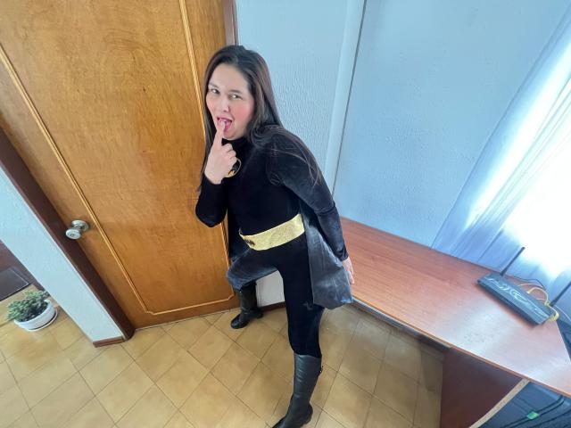 KarinaParker with dark hair, black outfit, grey boots, yellow belt, finger in mouth, standing by wooden door, wooden table with phone, and white curtain.