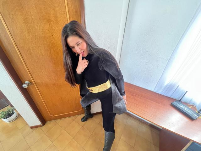 Karina Parker, com cabelos castanhos longos, vestindo roupa preta, um casaco cinza, um cinto amarelo e botas cinzas, lambendo o dedo, em pé perto de uma porta e janela de madeira, em um cômodo com piso de azulejos..