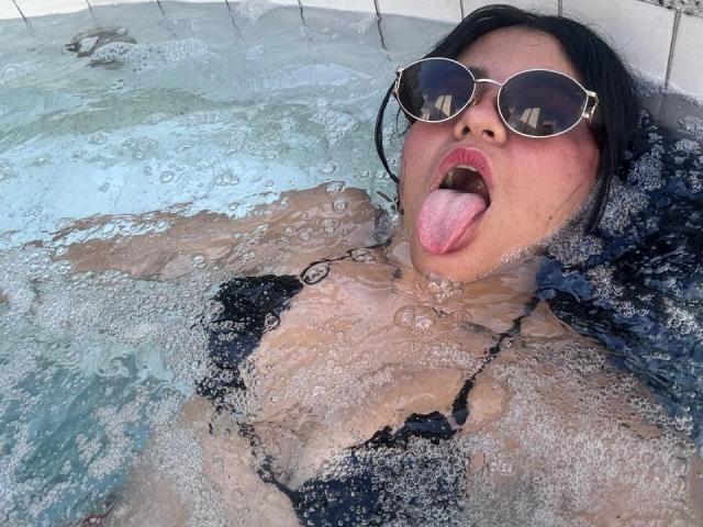 VALERY1CHAARMS in a pool, black bikini top, wet skin, tongue out, dark sunglasses, bubbles, water covering her chest, wet black hair, pink tongue, eyes closed.