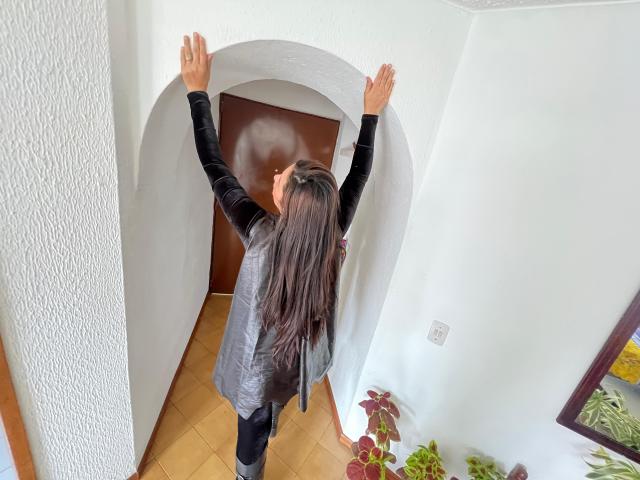 Karina Parker, com cabelos castanhos longos, blusa de veludo preta, calças legging pretas, braços levantados, tocando uma porta branca arqueada, porta marrom, paredes brancas, piso de madeira, pequena planta no canto inferior direito..