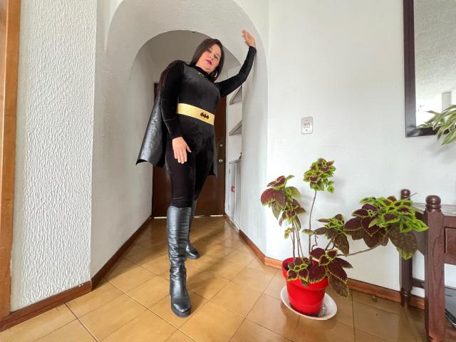 Karina Parker, con capelli castano scuro, indossa una giacca di pelliccia nera, una cintura gialla, pantaloni neri e stivali neri, appoggiata a un muro bianco, con un braccio alzato, in piedi vicino a una pianta in vaso rossa..