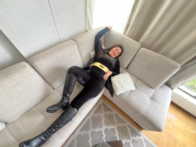Karina Parker está deitada em um sofá cinza, vestindo um conjunto preto com um cinto amarelo e botas pretas que chegam até o joelho, com um braço acima da cabeça e um travesseiro branco..