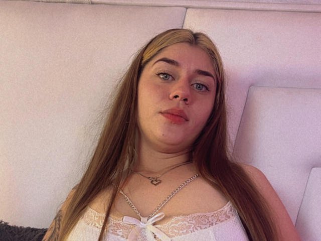 EllyAstown has long brown hair, blue eyes, and is wearing a white lace top with a bow. She's looking at the camera with a slight pout, necklace with heart.