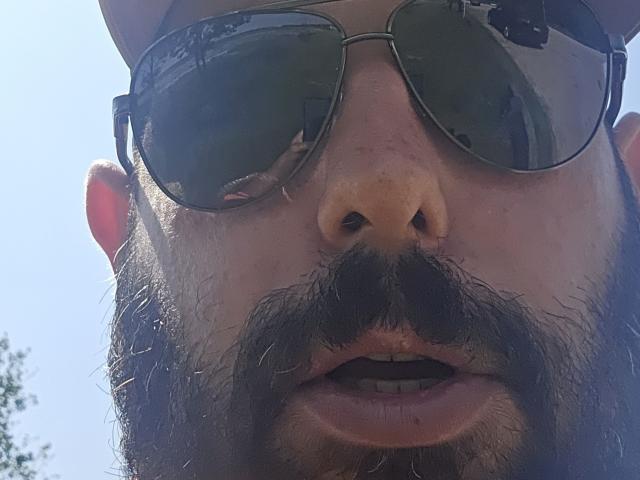 Erazion's face, with a thick black beard, wearing mirrored sunglasses, and a baseball cap, showing a slightly open mouth and a bit of sun reflection on the lenses.