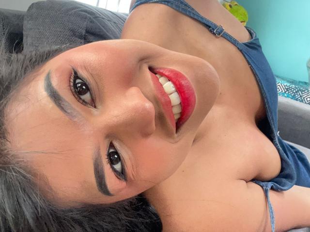 NairobiSwan with red lipstick, black eyebrows, and blue dress, lying down, smiling at camera, showing cleavage, black hair, and white teeth, looking cute and sexy.