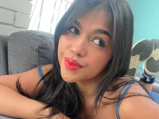 NairobiSwan, with dark straight hair and red lipstick, is lying on a grey couch, wearing a blue strap top, showing her cute face and smiling at the camera.