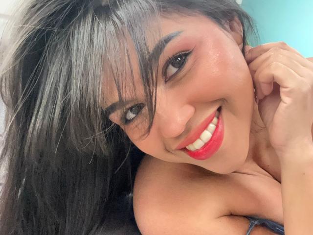 Nairobi: Una mujer con cabello negro largo, lápiz labial rojo y una gran sonrisa, mostrando sus senos y ojos brillantes, con una mano cerca de su mejilla, luciendo linda y sexy..