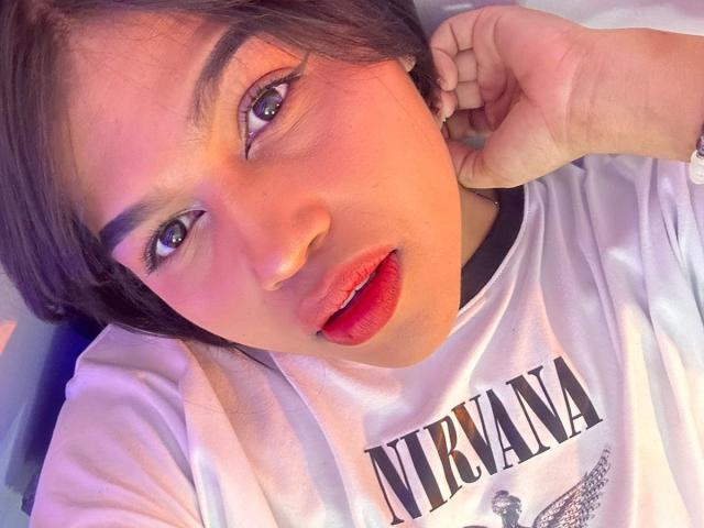NairobiSwan has red lips, brown eyes, and light skin. She's wearing a white shirt with "LIBYANA" and a small graphic. Her hand's near her ear. She looks up.