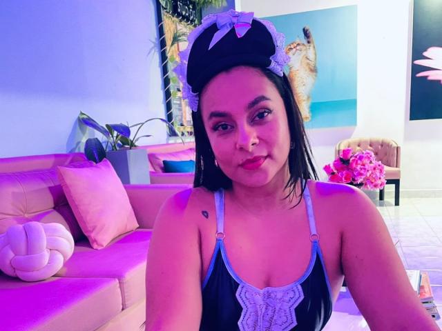 LunaSoler with dark hair, wearing a black and blue lace-trimmed top, and a white headband with a blue bow, sitting in a pink-lit room, smiling at the camera.