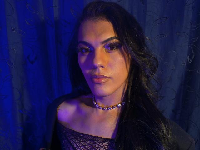 Ashley, una mujer con cabello negro largo, vestida con una blusa negra de encaje, un collar con adornos y iluminación azul. Tiene labios sensuales y carnosos, y pestañas gruesas y onduladas. Se pueden ver ligeramente sus senos..