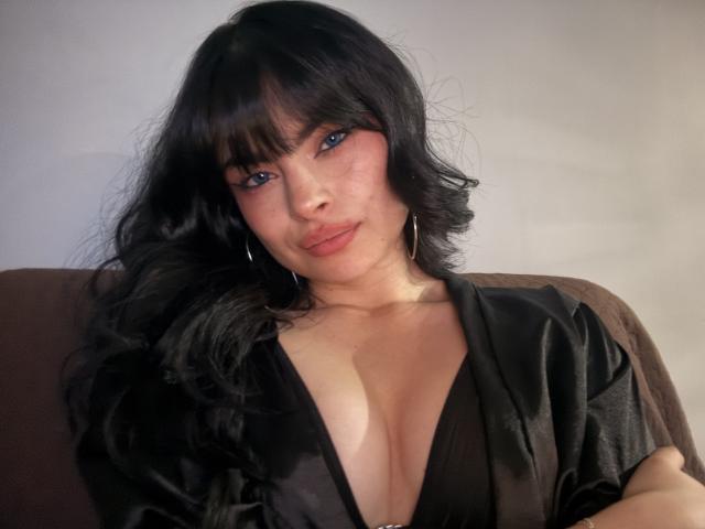 ClariceMontclair with black wavy hair, blue eyes, and pink lipstick, wearing a deep black V-neck dress, showing her tits. She's looking at the camera with a slight pout.