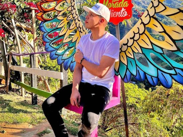 PelipeS with colorful angel wings, white cap, white shirt, black pants, sitting on pink chair, cross-arming, in sunny outdoor setting, looking to the side.