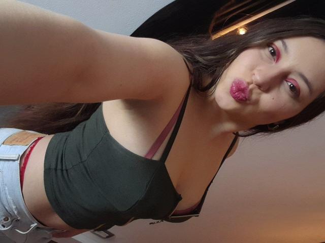 WidnyBeaumont, con lápiz labial rosa, labios fruncidos, un top corto verde oscuro, un sujetador negro, jeans blancos y ropa interior roja, mostrando sus pechos y glúteos, tomando un selfie..