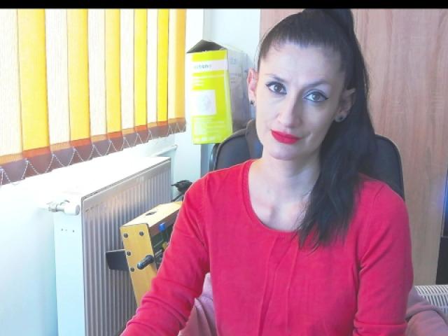 BeatrixCharmm has dark hair in a ponytail, wearing a red long-sleeve shirt, and red lipstick, sitting with a yellow box and radiator behind her.