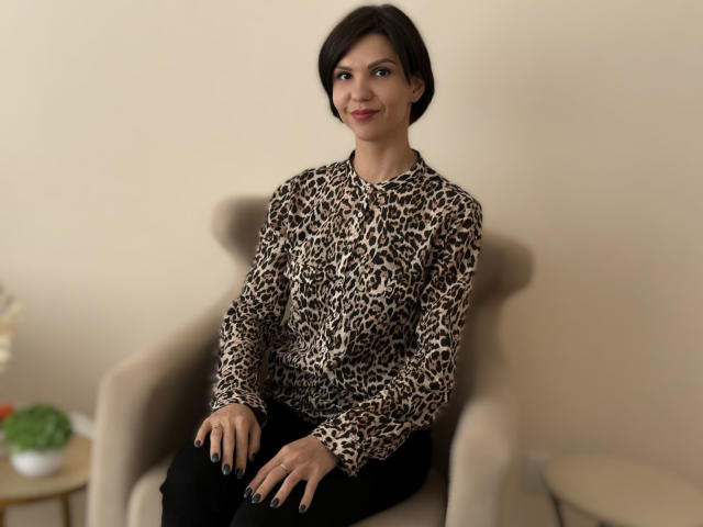 ElizabethSmile with short dark hair, wearing a leopard print shirt and black pants, sitting in a beige chair, smiling, black nails, small potted plant on left.