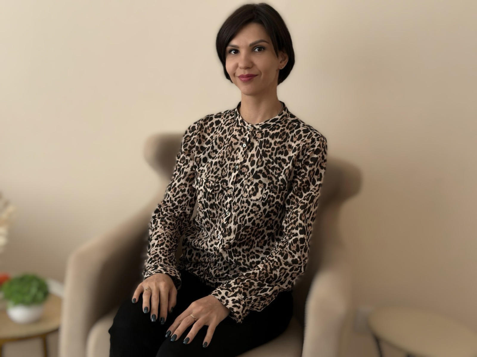 ElizabethSmile with short dark hair, wearing a leopard print shirt and black pants, sitting in a beige chair, smiling, black nails, small potted plant on left.