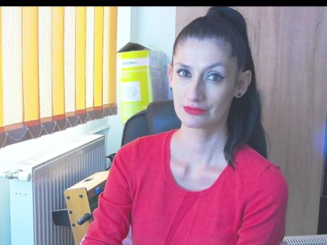 BeatrixCharmm with black hair in a ponytail, red lipstick, and a red long-sleeve shirt, sitting, smiling slightly, with a yellow box and vertical blinds in the background.
