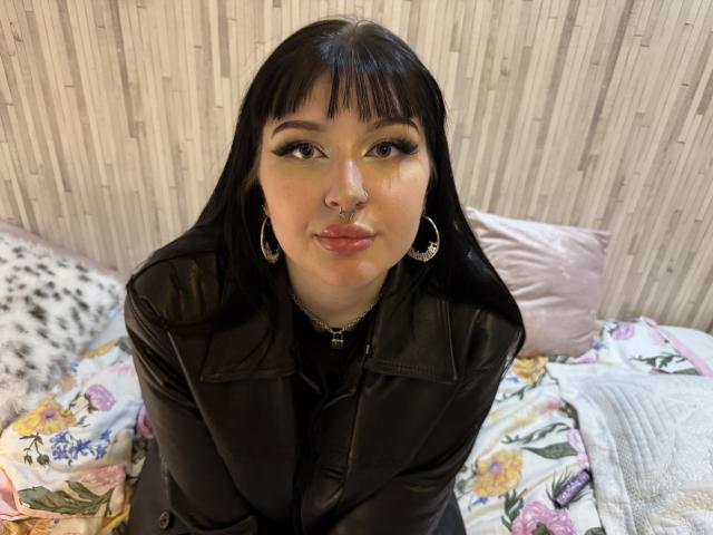 ShellyFire with black leather jacket, straight black hair, bangs, nose ring, hoop earrings, and lipstick, sitting on floral bed, looking up, smiling, medium tits, pale skin.