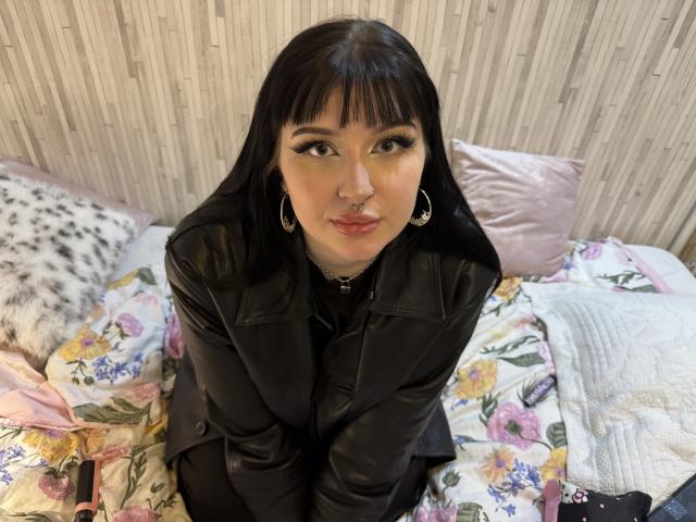 ShellyFire with black leather jacket, nose ring, and hoop earrings, sitting on a floral bed, looking up with pouty lips, black eyeliner, and long straight black hair.
