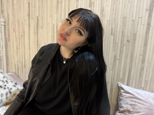 ShellyFire with long black hair, black leather jacket, black shirt, nose ring, and earrings, sitting with a slight tilt, wearing a small cross necklace, looking at the camera.