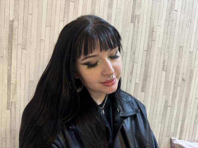 ShellyFire has black hair, eyeliner wings, nose ring, and lip ring, wearing a black leather jacket. She's smiling with long eyelashes and a cross necklace.