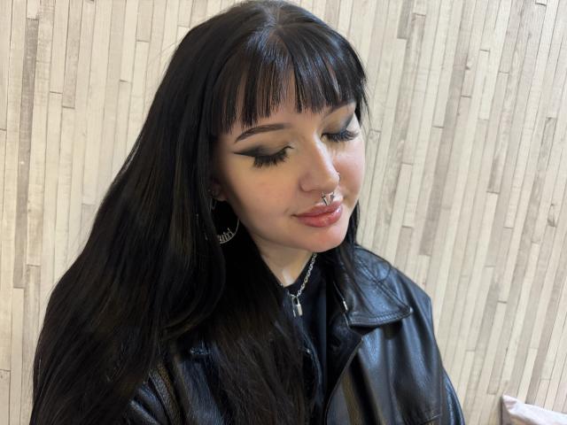 ShellyFire has long black hair, a nose ring, and eyeliner. She's wearing a black leather jacket and silver hoop earrings, with a cross necklace. Her lips are slightly pouty.