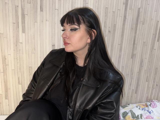 ShellyFire has black leather jacket, long straight black hair, bangs, nose ring, eyeliner, and black top, sitting with a pouty expression, wearing black pants, on a floral pillow.