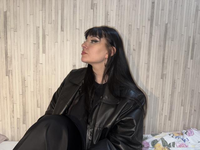 ShellyFire with black leather jacket, long straight black hair, and black pants, sitting with knees up, looking to the side, nose ring, and black eyeliner, on a bed.