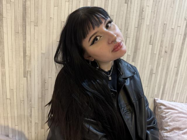 ShellyFire with long black hair, bangs, black eyeliner, nose ring, hoop earrings, leather jacket, and chain necklace, pouting with red lips, sitting on a beige pillow.