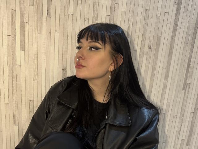ShellyFire with black leather jacket, straight black hair, bangs, eyeliner, nose ring, and earrings, sitting, looking to the side, wearing black shirt, in a photograph.