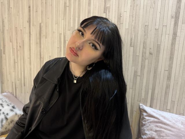 ShellyFire with long black hair, black leather jacket, black shirt, nose ring, hoop earrings, cross necklace, and bold eyeliner, looking up with a slight smile.
