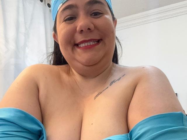 AndreaaX69, smiling, with bare tits, blue headscarf, and blue off-shoulder top, showing a small tattoo on her right shoulder, in a bright room.