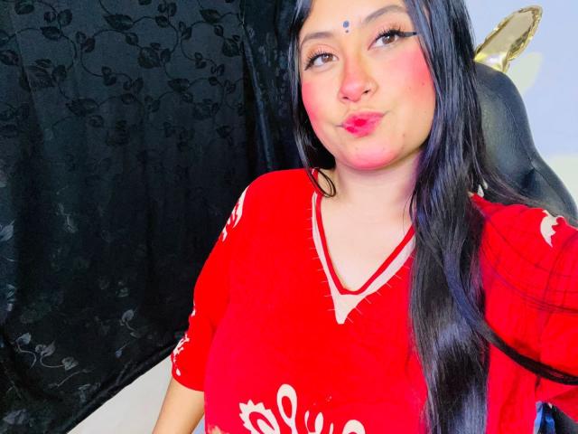 KalpanaAisha with long black hair, red dress, white floral patterns, and pink blush, pouting her full, red lips. She has a small bindi on her forehead.