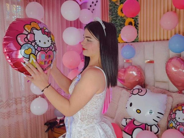 JadeNeferi with black hair, white lace dress, holding pink Hello Kitty balloon, surrounded by pink, white, and blue balloons, sitting on pink Hello Kitty pillow.