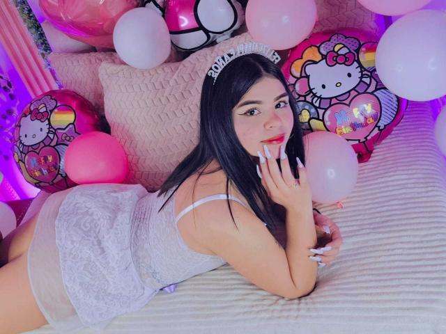 JadeNeferi, con cabello largo y negro, vestida con un vestido blanco de encaje y una tiara, está acostada en una cama, sosteniendo un globo blanco, mostrando sus glúteos, y rodeada de globos de Hello Kitty..