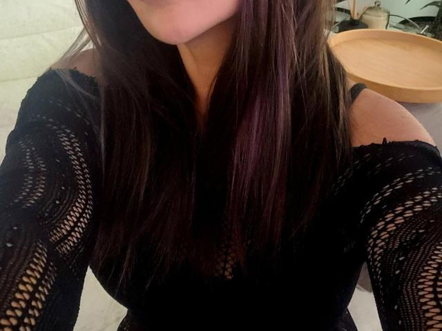 EmmaNlovely with long brown hair, wearing a black lace off-shoulder top, showing her boobs through the intricate pattern. Her face is partially visible, upper body shot.